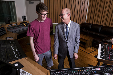 A man in a suit jacket and tie smiles at another man in a t-shirt to his right in a music studio