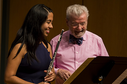 A man with white hair smiles while looking down as a woman to his right is holding a flute