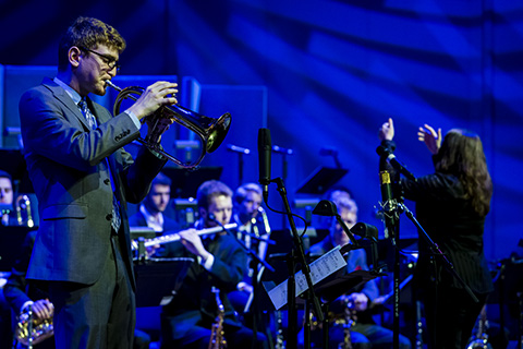 A trumpet player from the FROST Studio Jazz Band is playing while a conductor directs a band in the background
